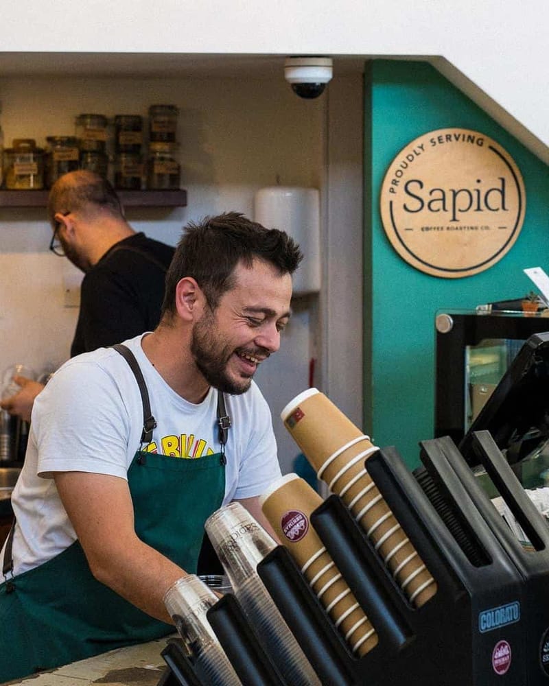 A smiling barista in a green apron behind the counter, a Proudly serving Sapid sign on the teal wall.