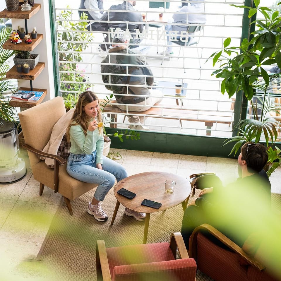 Inside Seniman: two friends with coffee in armchairs by the green-framed window, plants all around, people at the tables outside.