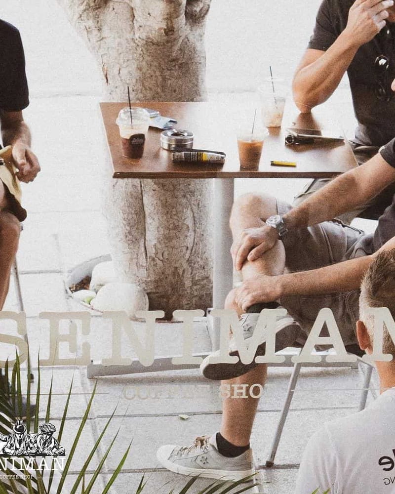 Regulars around an outside table with iced coffees under a tree on the pavement.